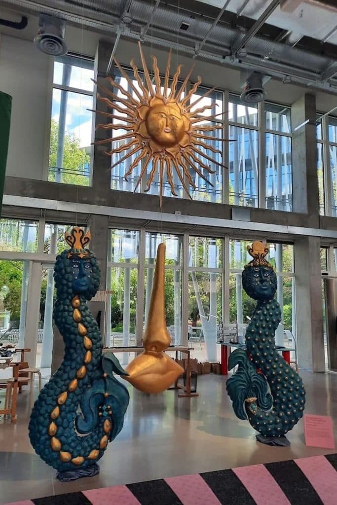Colorful handcrafted sculptures, including two blue figures and a golden sun, displayed inside the bright Galerie du 19M in Paris.
