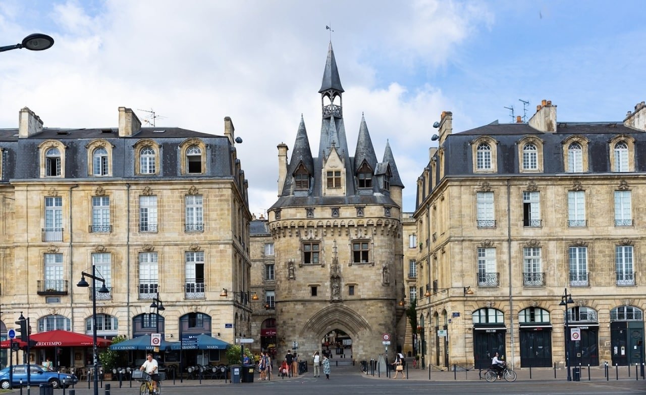 The historic Porte Cailhau, a stunning medieval gateway with pointed turrets and intricate stonework, stands between elegant limestone buildings in Bordeaux’s city center, a must-see landmark often recommended in Paris travel blog guides for those exploring France beyond the capital.