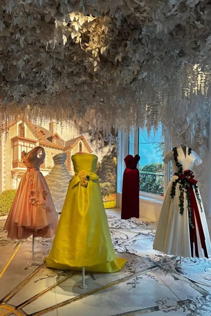 Colorful Dior couture gowns displayed beneath a floral ceiling at La Galerie Dior in Paris.