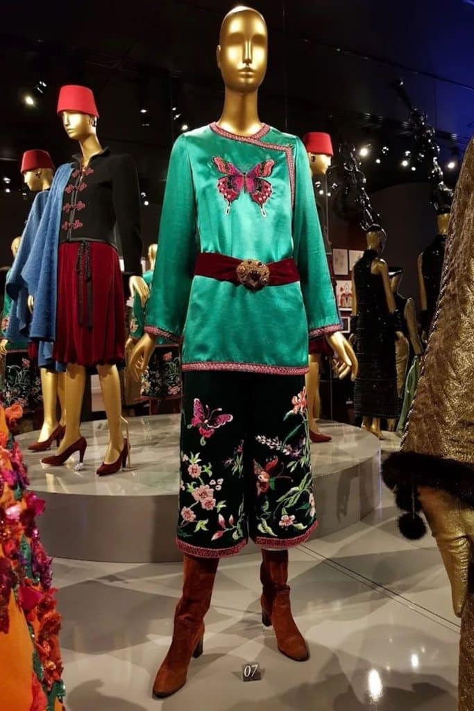 Mannequin wearing a turquoise embroidered top, floral trousers, and red boots in a colorful Yves Saint Laurent fashion exhibition.