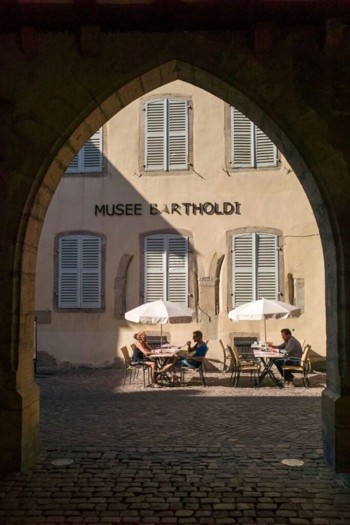 A charming courtyard café outside the Bartholdi Museum in Colmar, France, viewed through a stone archway. Visitors sit under white umbrellas, enjoying the tranquil atmosphere of this historic site dedicated to Frédéric Auguste Bartholdi, the sculptor of the Statue of Liberty. A must-visit stop when spending one day in Colmar.