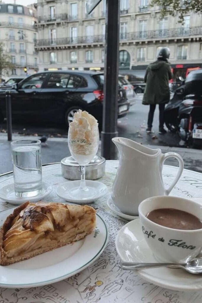 Cozy outdoor café table in Paris with a cup of the best hot chocolate in Paris at Café de Flore, accompanied by a slice of apple tart, whipped cream in a glass, and a small pitcher, set against a classic Parisian street scene.