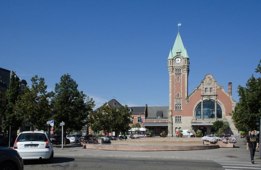 The historic Gare de Colmar (Colmar Train Station) with its striking red brick façade, tall clock tower, and green copper roof, set against a clear blue sky. The surrounding plaza is lined with trees, flowers, and cars, making it a central arrival point for travelers spending one day in Colmar.