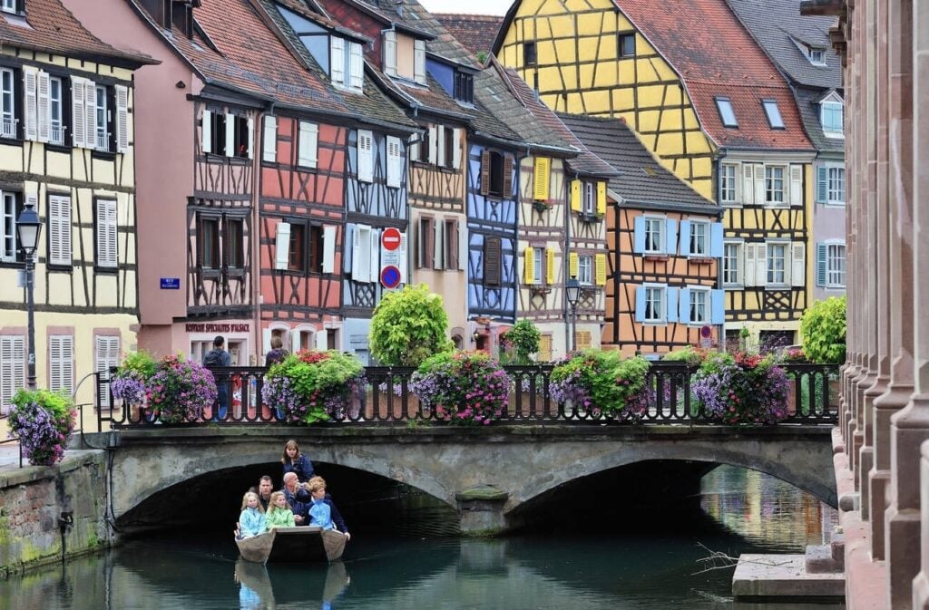 A scenic boat ride through Little Venice in Colmar, France, with tourists gliding along the canal under a charming stone bridge adorned with vibrant flowers. The backdrop features colorful half-timbered houses, creating a fairy-tale atmosphere. A must-do experience when spending one day in Colmar to soak in the beauty of Alsace.