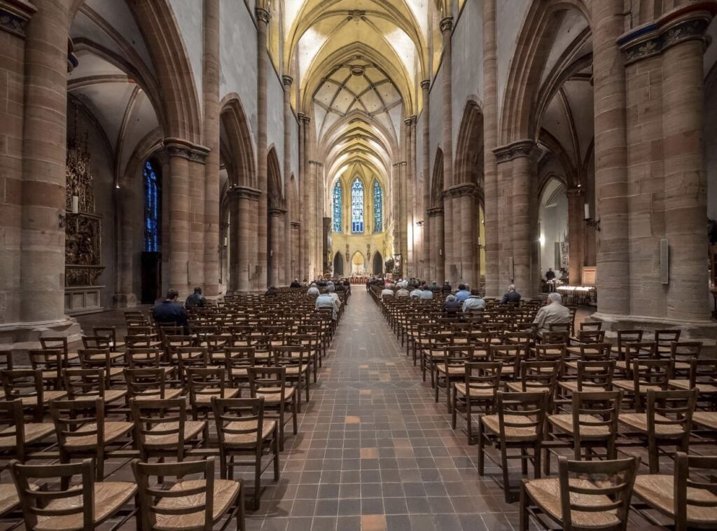 The grand interior of St. Martin’s Church in Colmar, France, showcasing its soaring Gothic arches, intricate stone columns, and stunning stained-glass windows. Rows of wooden chairs line the nave, leading toward the illuminated altar. A must-visit landmark when spending one day in Colmar to admire its historic architecture.