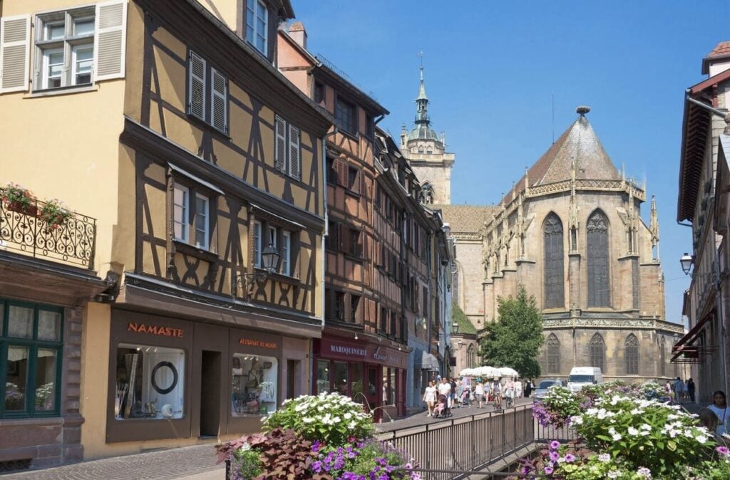 A picturesque street in Colmar, France, leading to St. Martin's Church, a stunning Gothic cathedral with intricate architecture. Half-timbered buildings, boutique shops, and vibrant flower arrangements line the cobblestone path, capturing the essence of Alsace. A must-see landmark when spending one day in Colmar.