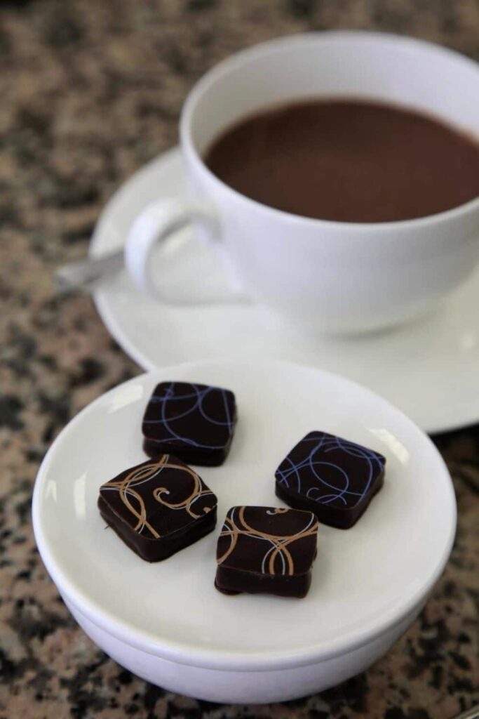 Close-up of four elegantly decorated square chocolates on a white plate, placed in front of a white cup filled with the best hot chocolate in Paris at Jacques Genin, highlighting the rich, velvety texture of the drink and the artisanal quality of the chocolates on a granite tabletop.