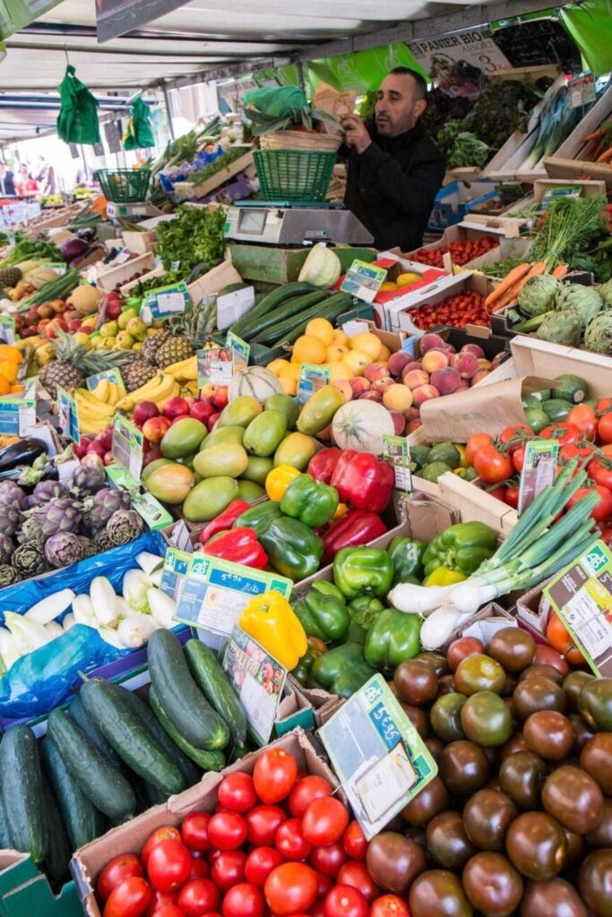 A vibrant stall at Marché d'Aligre, a flea markets in Paris displays an abundance of fresh fruits and vegetables, with a vendor weighing produce for a customer.