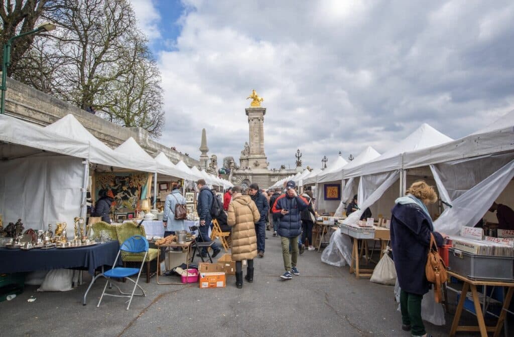 Outdoor flea market near Pont Alexandre III in Paris, featuring rows of white tents filled with vintage treasures, vinyl records, antiques, and art. Locals and tourists browse under cloudy skies, with the golden statue of Fame in the background adding a classic Parisian flair to the vibrant flea market atmosphere.