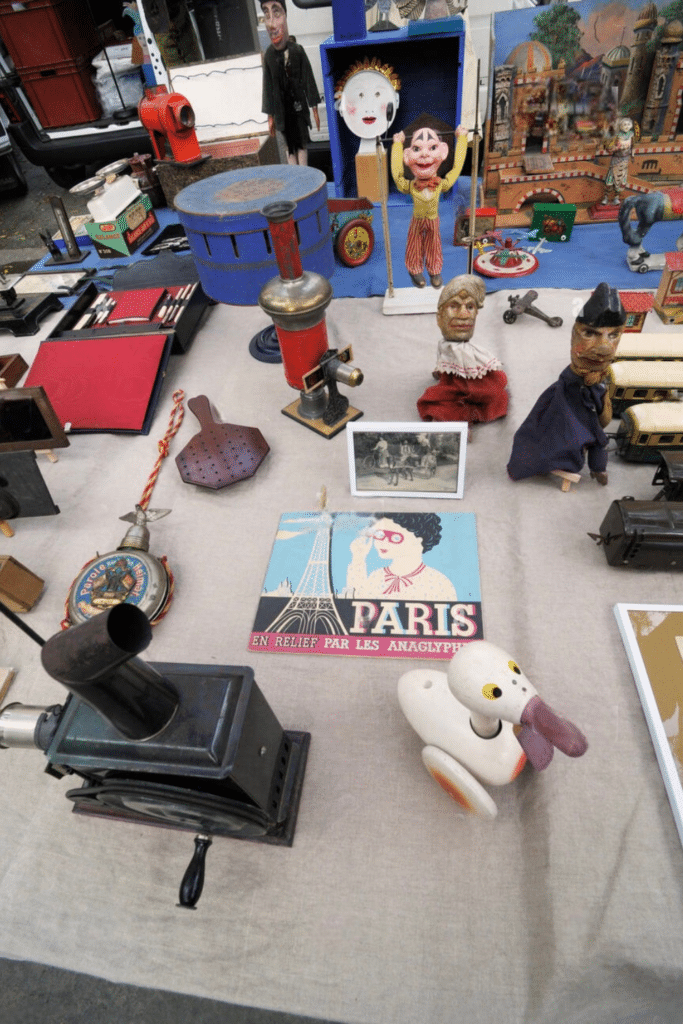 A table at a flea markets in Paris is filled with an assortment of vintage collectibles, including old-fashioned puppets, antique optical devices, a mechanical duck toy, and a colorful "Paris" sign, all arranged on a beige cloth alongside nostalgic trinkets and decorative items.