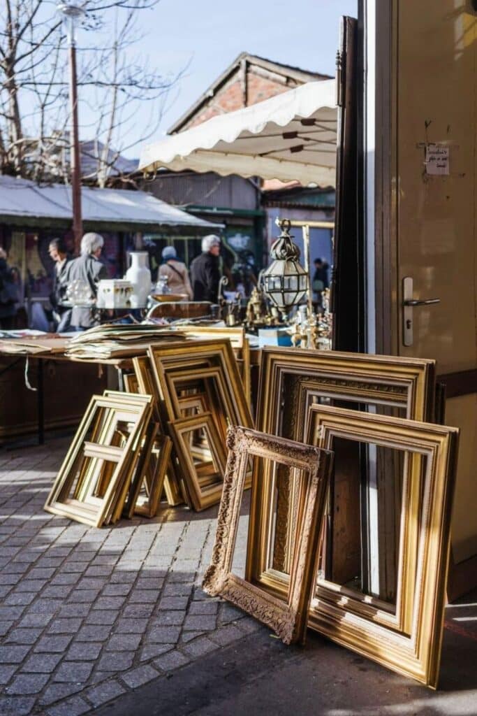 A collection of ornate vintage gold frames leaning against a wall at Marché aux Puces de Saint-Ouen, one of the most iconic flea markets in Paris. The scene captures the charm of treasure hunting in Paris, with antique chandeliers, ceramics, and curious shoppers browsing under white awnings on a sunny day.
