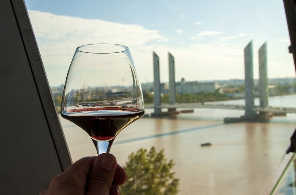 A hand holds a glass of red wine against the backdrop of the Pont Jacques Chaban-Delmas in Bordeaux, France, showcasing one of the top things to do in Bordeaux—enjoying local wine with a scenic view.