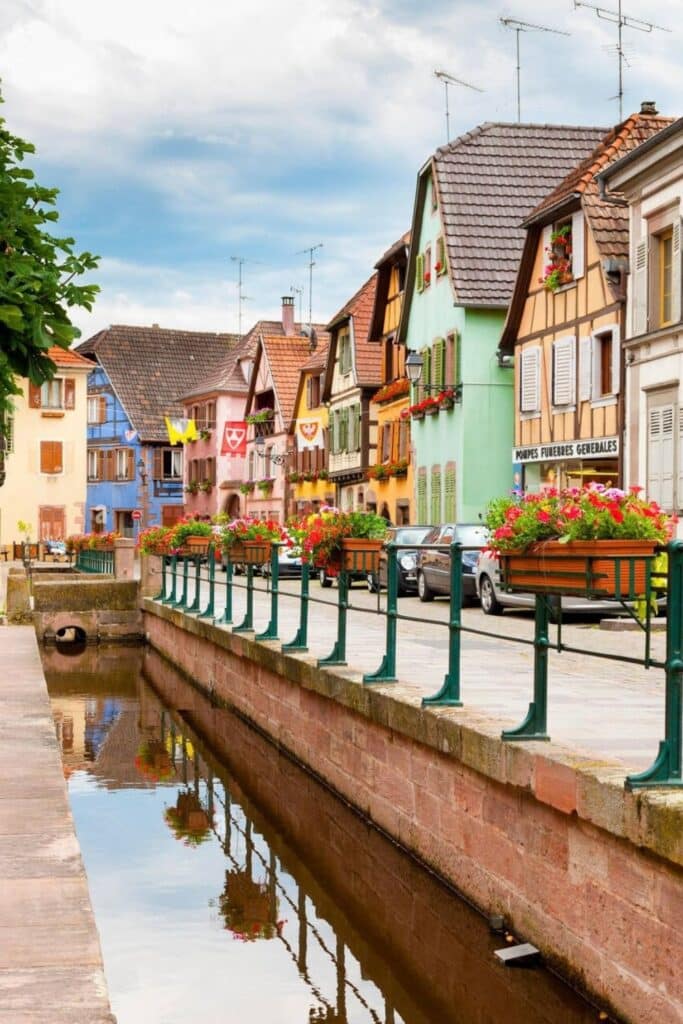 Colorful houses along the canal in Huningue, a charming place to visit in Alsace, with vibrant flowers adorning the windows and picturesque reflections in the water.