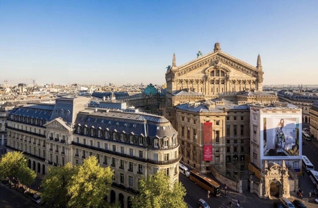 An aerial view of the 9th arrondissement in Paris, featuring the ornate Palais Garnier opera house and surrounding Haussmann-style buildings under a clear blue sky.