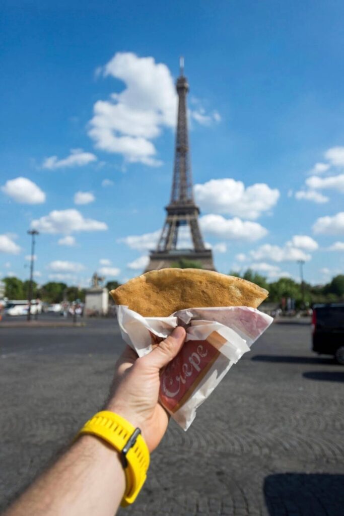 A close-up of a hand wearing a yellow watch, holding a warm crêpe wrapped in paper that reads "Crêpe," with the Eiffel Tower rising in the background under a bright blue sky—highlighting a quintessential Best Crêpes in Paris moment.