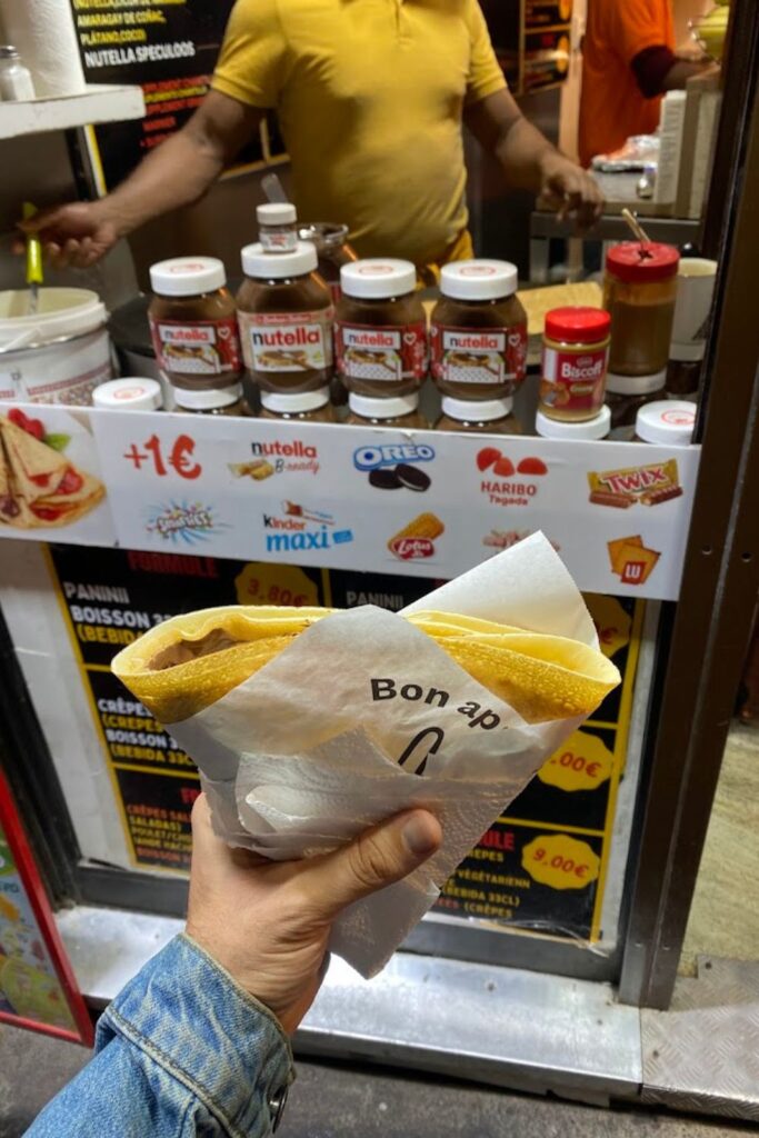 A freshly made Nutella crêpe wrapped in paper, held in front of a bustling Parisian street vendor, showcasing the best crêpes in Paris.