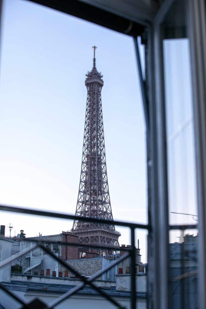 A close-up view of the Eiffel Tower framed by an open window, showcasing the charm of Paris hotels with Eiffel Tower views.