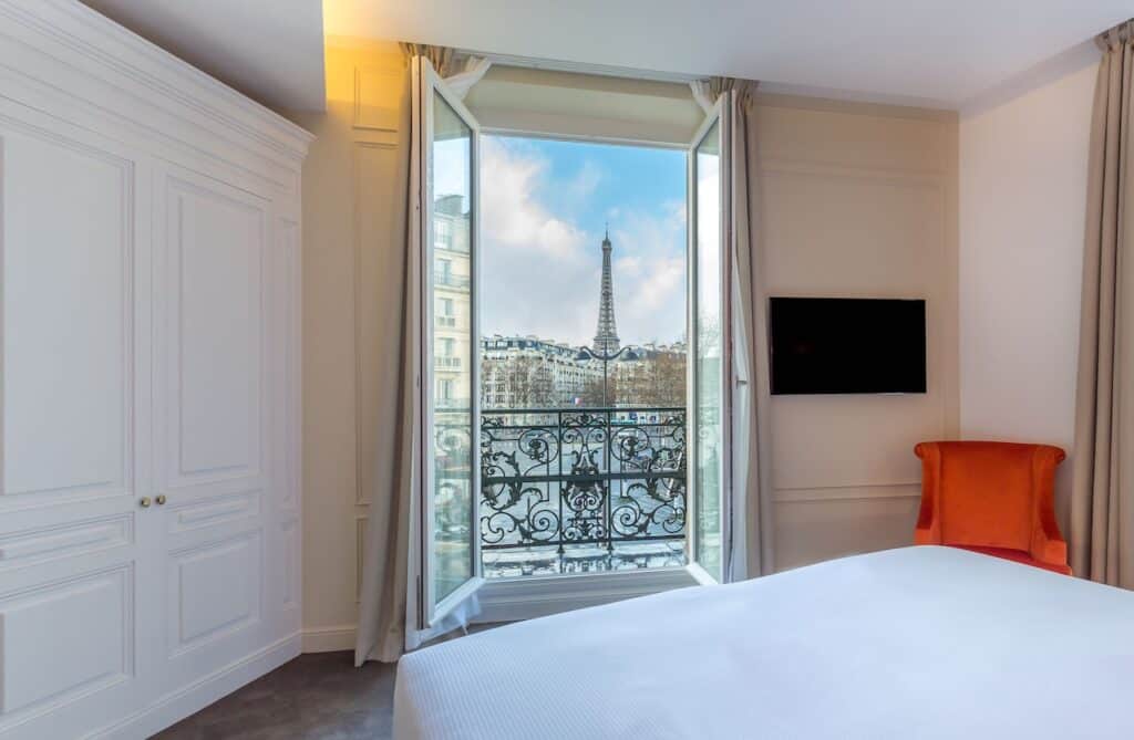 A bright hotel room featuring a balcony with ornate iron railings and a stunning Eiffel Tower view, ideal for Paris hotels with Eiffel Tower views.