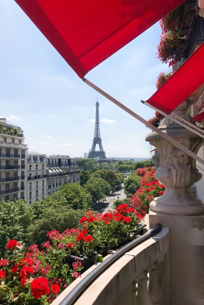 A picturesque balcony adorned with red flowers and awnings, offering a stunning view of the Eiffel Tower, perfect for Paris hotels with Eiffel Tower views.