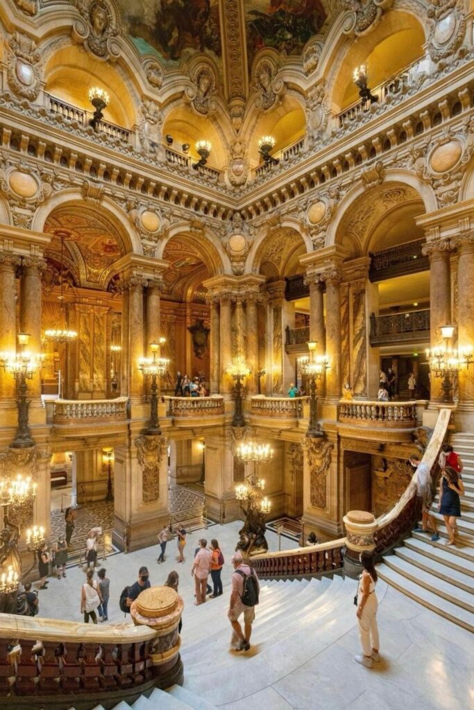 The grand staircase inside the Palais Garnier in the 9th arrondissement of Paris, featuring ornate marble, gilded details, chandeliers, and intricate ceiling murals as visitors explore the opulent interior.