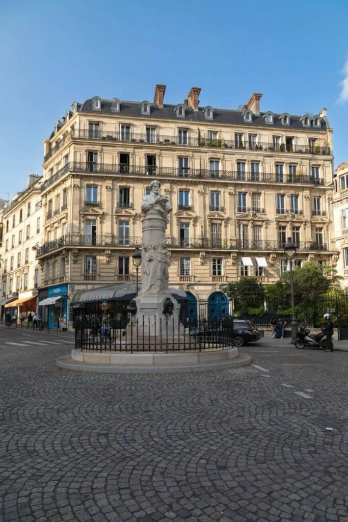 A cobblestone square in the 9th arrondissement of Paris, featuring a central stone monument surrounded by a wrought iron fence, with elegant Haussmann-style buildings in the background.