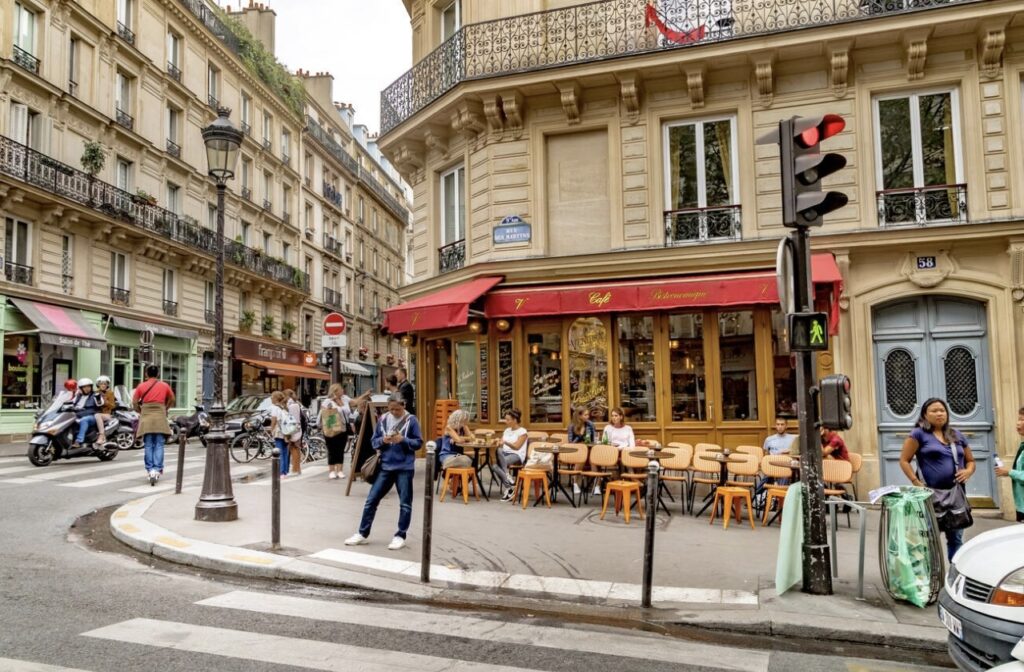 A bustling street corner in the 9th arrondissement of Paris, featuring a charming café with a red awning, outdoor seating, and nearby pedestrians amidst classic Haussmann-style buildings.