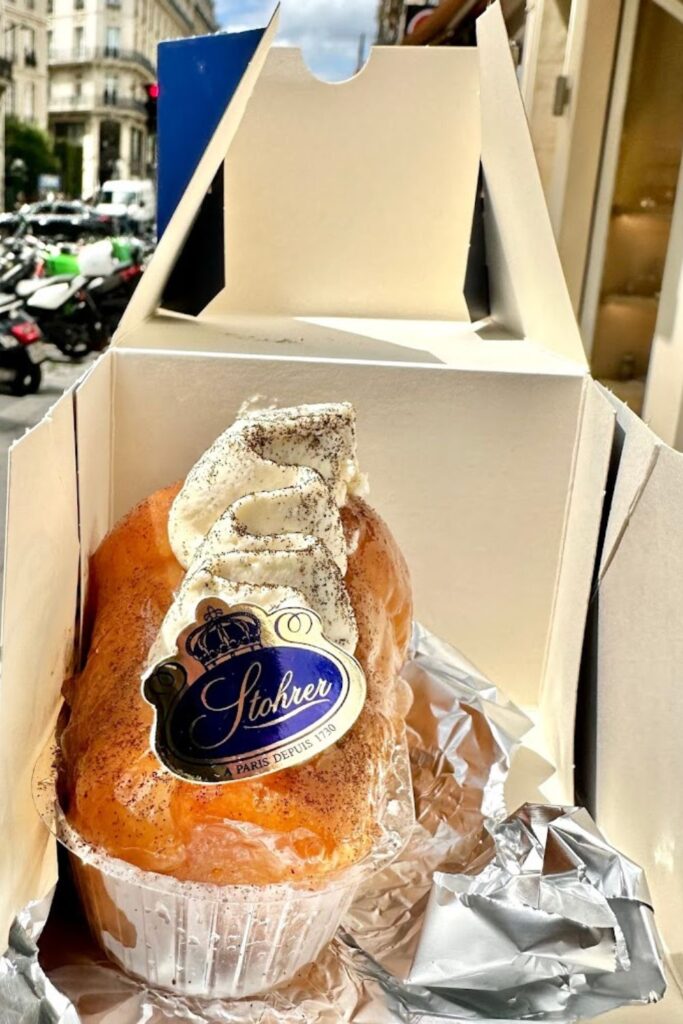 A rum baba dessert topped with whipped cream from the historic Stohrer patisserie, displayed in its box with the streets of the 9th arrondissement of Paris visible in the background.