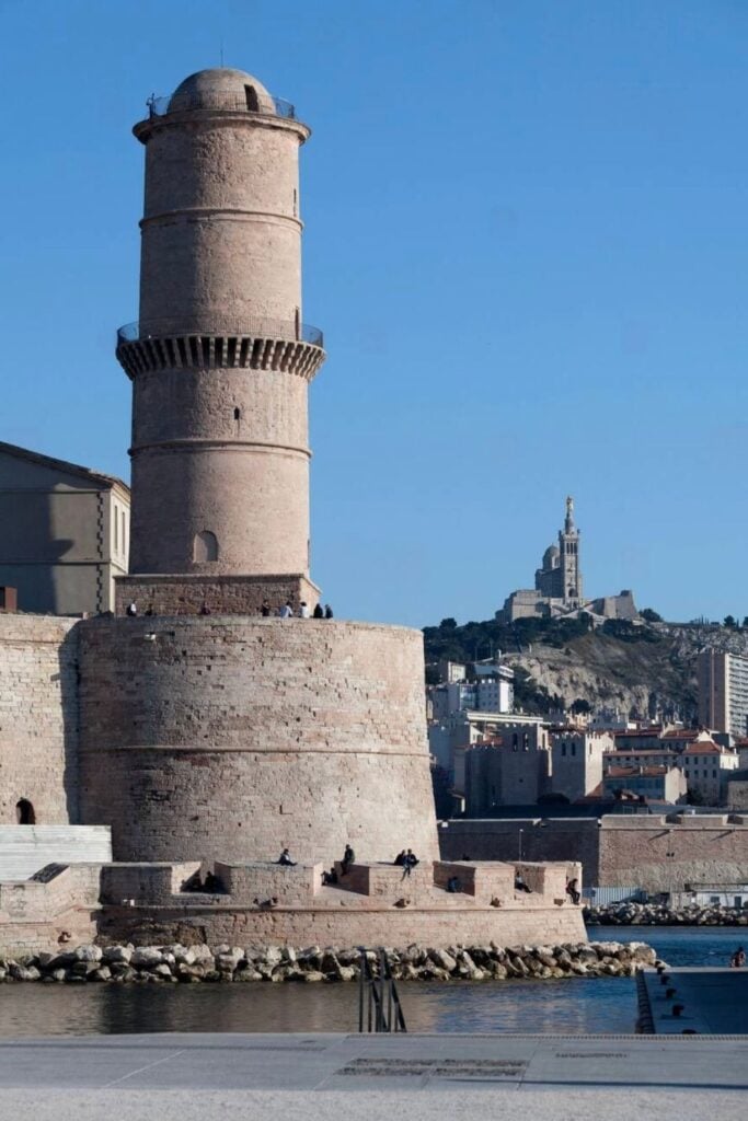 The historic Fort Saint-Jean, with its cylindrical stone tower overlooking the waterfront, is a must-visit landmark and one of the top things to do in Marseille France, with Notre-Dame de la Garde visible in the background.
