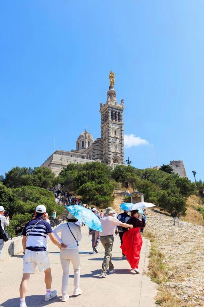 Visitors walk up the sunlit path to Notre-Dame de la Garde, a stunning basilica with a golden statue overlooking the city, making it one of the top things to do in Marseille France.