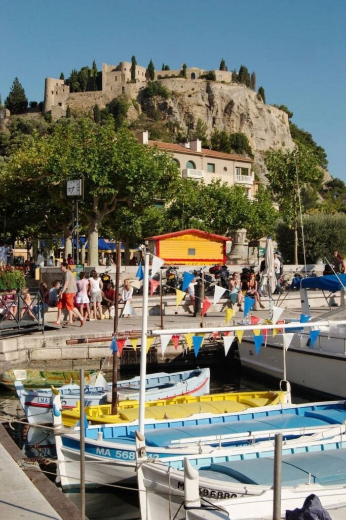 Colorful fishing boats line the lively waterfront of a Provençal village near Marseille, with a medieval hilltop castle in the background—exploring nearby coastal towns is one of the best things to do in Marseille France.