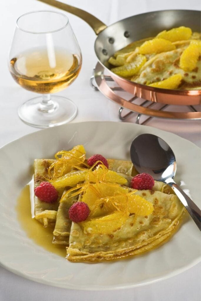 A plate of Crêpes Suzette, a classic traditional French foods, served with caramelized orange sauce, raspberries, and spun sugar, alongside a glass of brandy.
