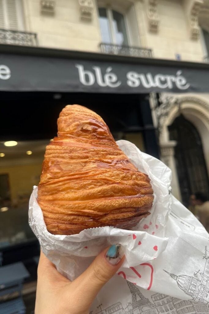 A beautifully layered and golden-brown croissant, a staple of traditional French foods, held in front of the famous Blé Sucré bakery in Paris.