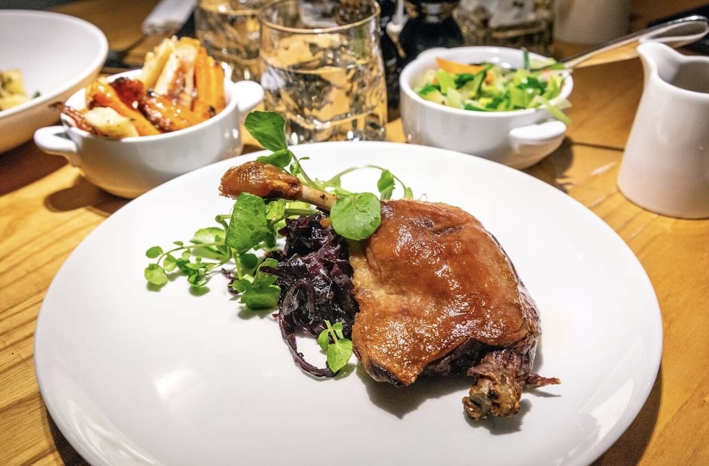A plate of crispy duck confit, a classic traditional French foods, served with fresh greens and braised red cabbage, with side dishes in the background.