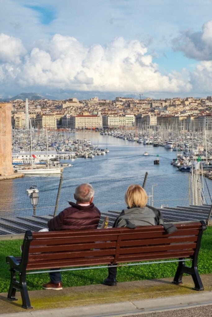 A couple sits on a bench overlooking Marseille’s Old Port, admiring the yachts, historic buildings, and distant mountains—soaking in this panoramic view is one of the most relaxing things to do in Marseille France.