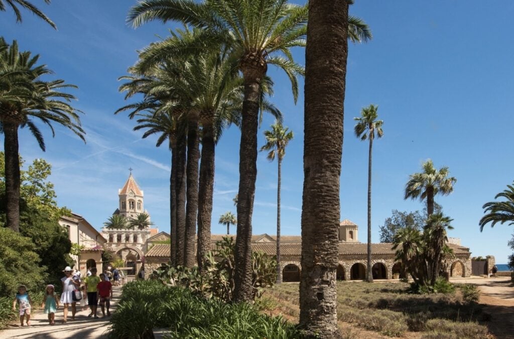 Families stroll through palm-lined paths toward the historic abbey on Saint-Honorat Island, a serene option for day trips from Nice, France.