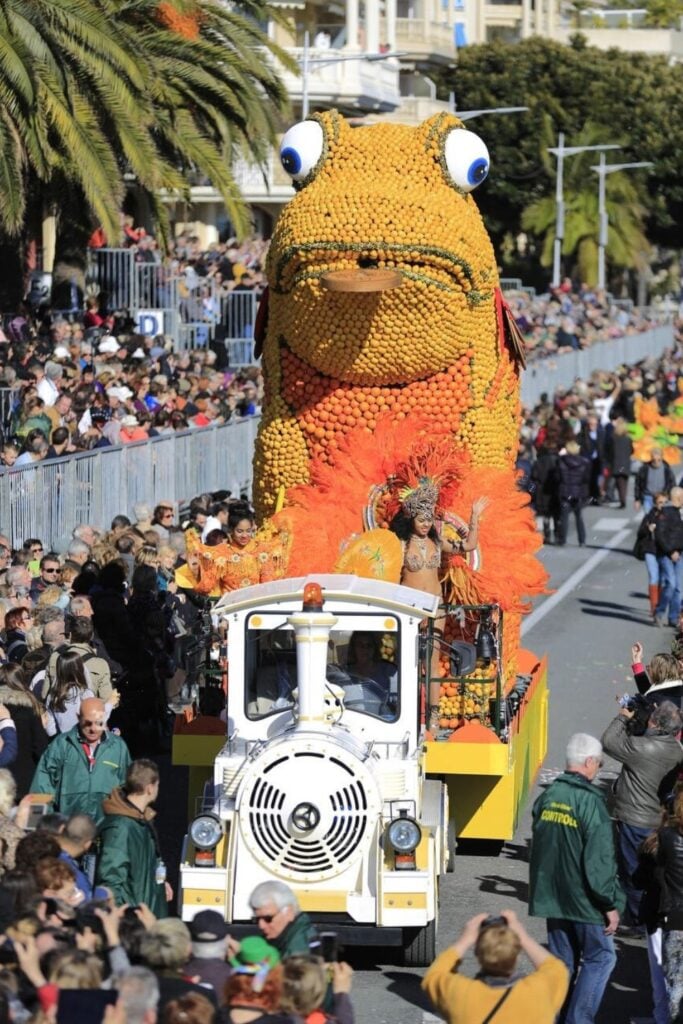 A vibrant parade float featuring a giant citrus-covered frog and performers in bright orange costumes entertains crowds at the Menton Lemon Festival, one of the most exciting things to do in Menton France.