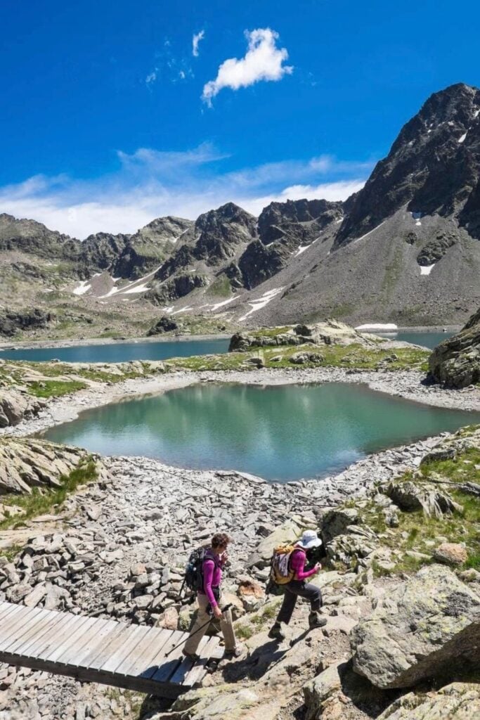 Two hikers cross a rocky path by a serene alpine lake in the Mercantour National Park, a stunning destination for day trips from Nice, France.