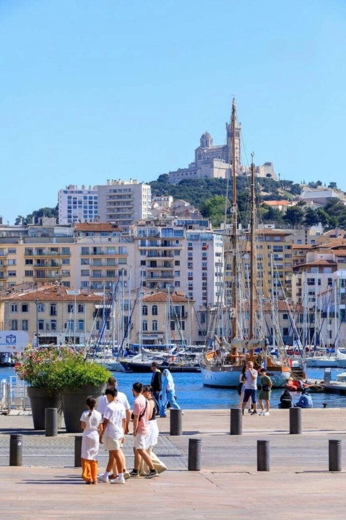 People stroll along Marseille’s Old Port, with sailboats in the harbor and the iconic Notre-Dame de la Garde basilica perched on the hill—exploring this vibrant waterfront is one of the top things to do in Marseille France.