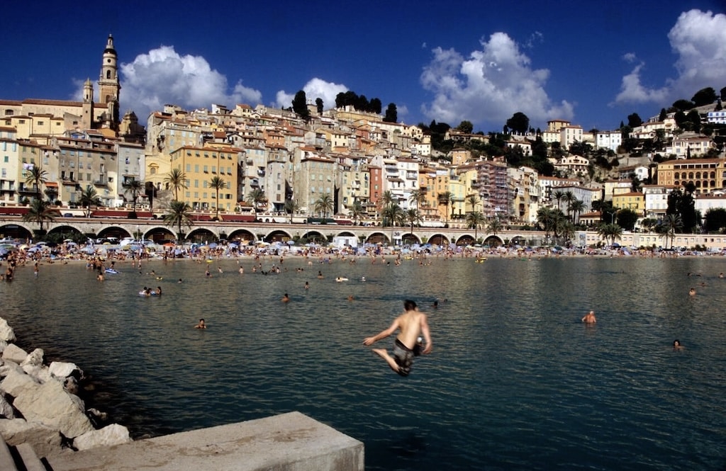 A lively beach scene in Menton, France, with people swimming in the clear blue water and a young man mid-dive, set against the backdrop of colorful hillside buildings—one of the top things to do in Menton France.