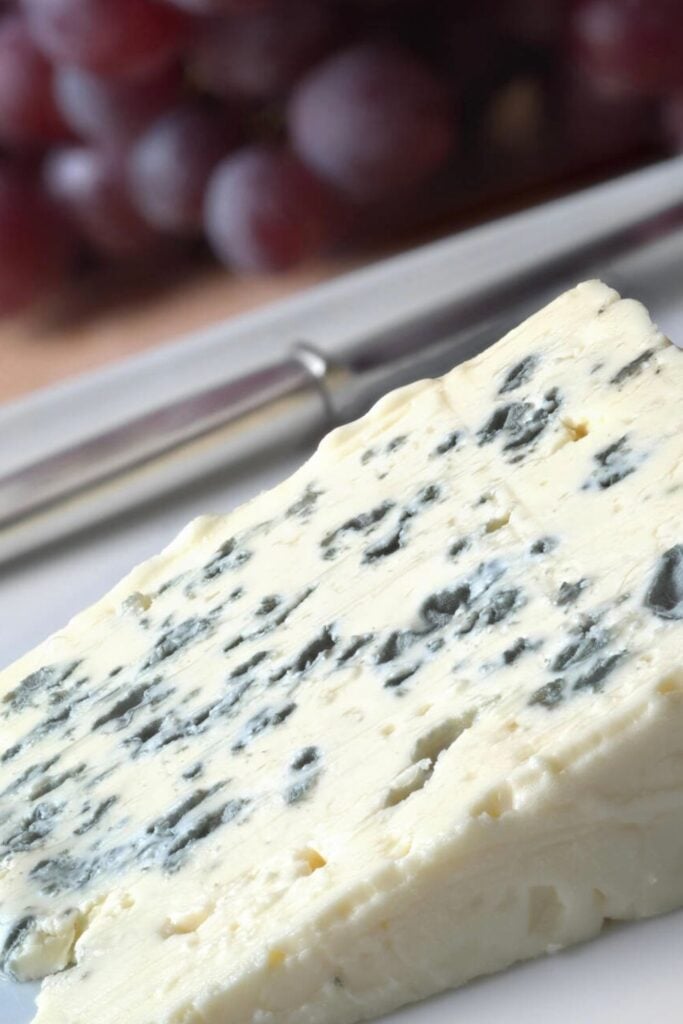 A creamy wedge of blue-veined Roquefort cheese, a bold and flavorful staple of traditional French foods, served with grapes in the background.