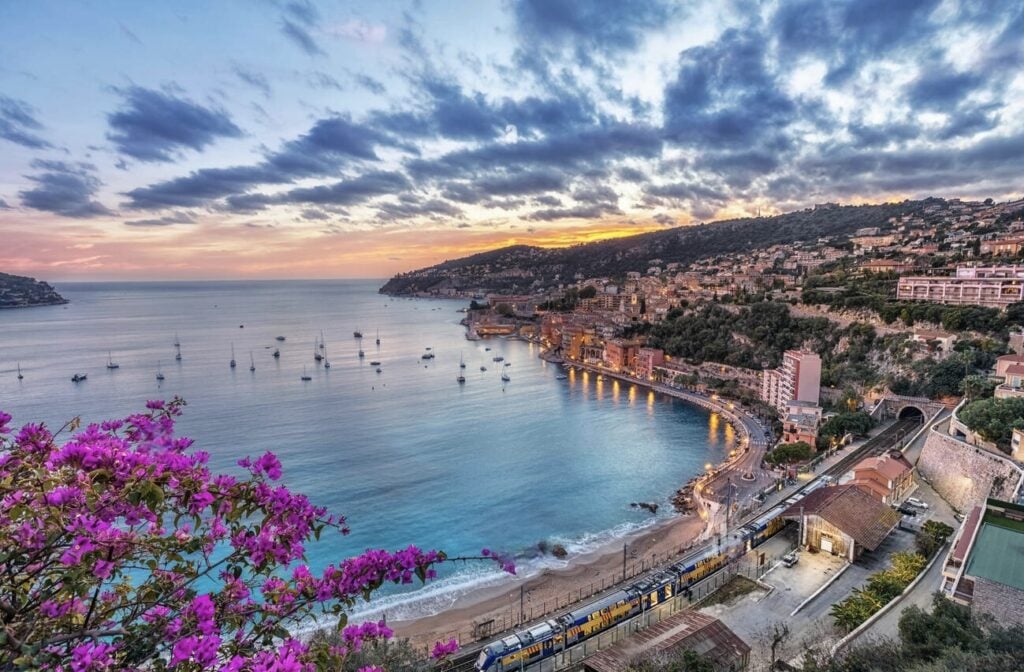 The bay of Villefranche-sur-Mer glows under a vibrant sunset, with sailboats dotting the calm waters and a coastal train arriving, making it a breathtaking option for day trips from Nice, France.