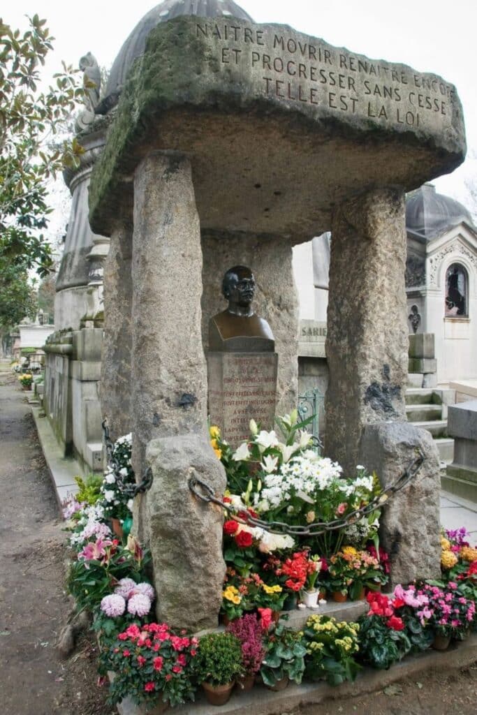 A stone monument at Père Lachaise Cemetery features a bronze bust beneath a large inscribed slab, supported by rough-hewn pillars, with chains draped across the front and surrounded by vibrant flowers.
