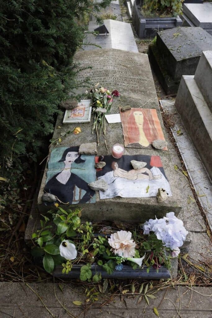 The grave at Père Lachaise Cemetery is adorned with flowers, candles, and artistic tributes, including paintings inspired by the distinctive portrait style of Amedeo Modigliani.