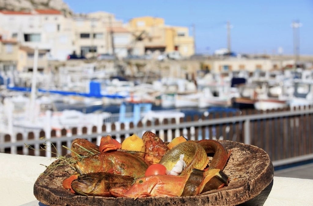 A rustic platter of fresh Mediterranean fish, tomatoes, garlic, and herbs, set against a scenic Marseille harbor—a must-try dish in any Marseille Food Guide.