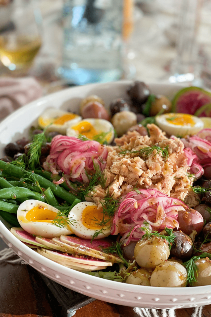 A vibrant cuisine Niçoise inspired salad with flaky tuna, jammy eggs, pickled red onions, potatoes, green beans, olives, and fresh dill in a white serving bowl.