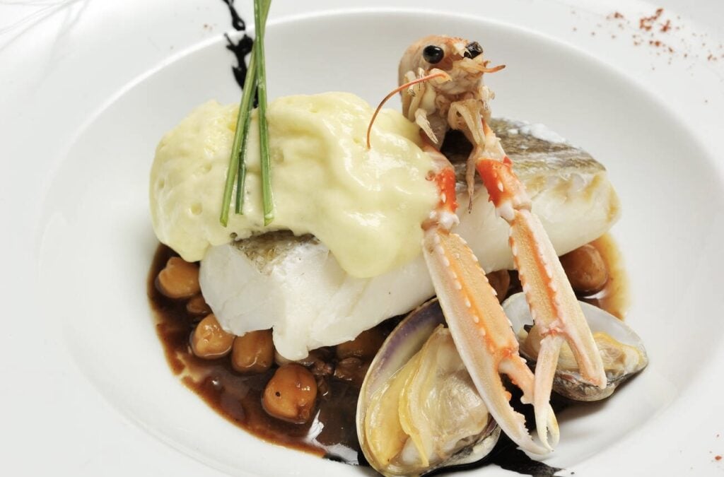 A refined seafood dish featuring cod with a creamy sauce, chickpeas, clams, and a langoustine garnish—an exquisite find in any Marseille Food Guide.