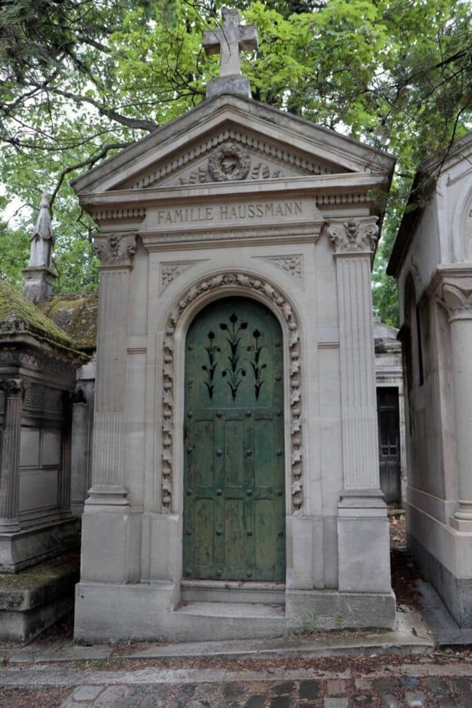 The Haussmann family mausoleum at Père Lachaise Cemetery is an elegant stone structure with intricate carvings, a weathered green door, and a cross-topped pediment nestled among lush trees.