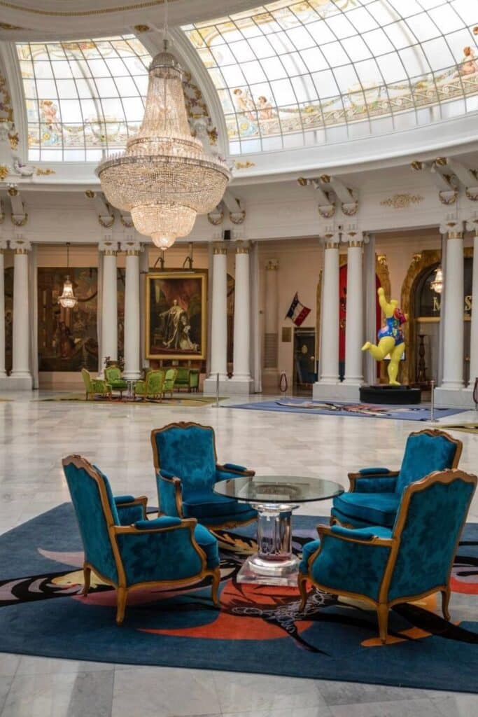 The grand interior of the Musée Masséna in Nice showcases an opulent chandelier, elegant blue velvet chairs, classical artwork, and a stunning glass-domed ceiling—offering a glimpse into history and one of the must-see things to do in Nice.