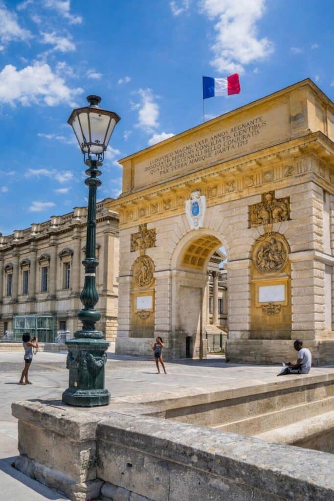A historic stone arch in Montpellier, France, adorned with intricate carvings and a French flag, stands under a bright blue sky, with tourists exploring the area—one of the many things to do in Montpellier.
