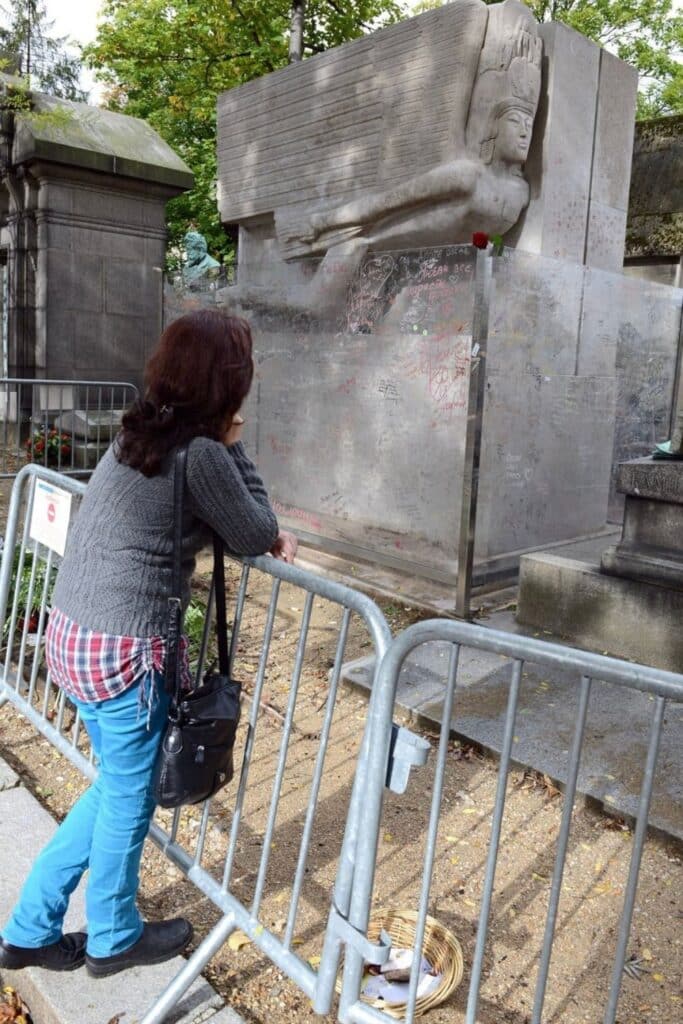 A visitor leans on a metal barrier, gazing at Oscar Wilde's tomb in Père lachaise cemetery which features a striking winged stone sculpture and a glass screen covered in lipstick marks and messages from admirers.